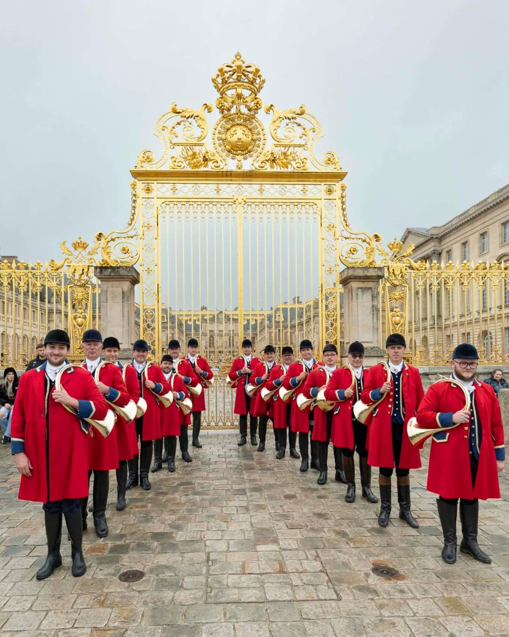 Les sonneurs du Débuché de Paris en habit rouge, alignés devant la grille dorée du château de Versailles